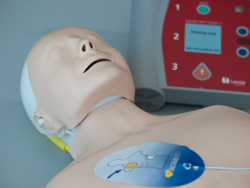 A CPR manikin with an AED on the floor close by. An AED pad is stuck to the manikin's chest under the right shoulder.