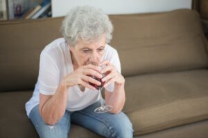 An older woman wearing jeans and a white t-shirt, sits on a beige sofa drinking a glass of red wine and looking depressed.