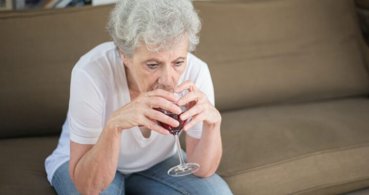 An older woman wearing jeans and a white t-shirt, sits on a beige sofa drinking a glass of red wine and looking depressed.