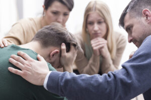 Close-up of a devastated young man holding his head in his hands and a group of friends in a supportive pose around him